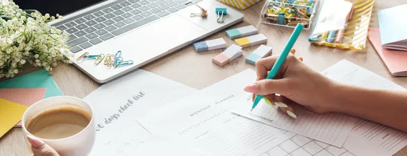 Office desk with laptop, sheets of paper, erasers and a womans hand holding a pen and cup of coffee.